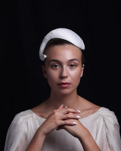 The model is showcasing an “S” shaped sateen bridal headband. The piece starts from the right side and curves upwards head-hugging the head while getting thicker.  The artistic headband is perfect for modern brides.