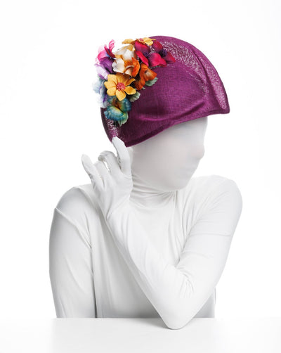 The hat the model wears in the picture is a striking purple headpiece with a unique sculpted shape. It is adorned with a vibrant array of colourful flowers on one side, featuring shades of yellow, red, pink, and orange, along with some green leaves. 