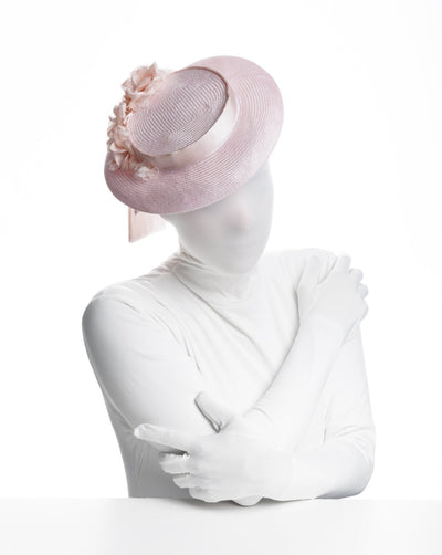 The Model is wearing a small-sized boater-style pink parasisal hat, while the hat covers the head with a smaller crown and shorter brim. The crown of the hat is rounded with a pink silk sateen belt, and towards the right to the back, a mixture of pink flowers of different sizes and kinds. You can see the extra belt of the hat falling off to the hat at the back.