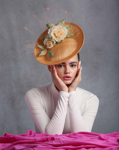 The image shows a woman wearing a stylish fascinator hat tilted to the side. The hat is a warm peach colour and adorned with two large, realistic-looking cream-coloured roses with a touch of peach and green leaves. A delicate, light peach-coloured netting with small decorative accents is attached to the hat, adding an elegant and whimsical touch. The saucer-style hat has an upward curve from the centre that goes higher in the back.  The hat is worn, slightly tilted to the right side.