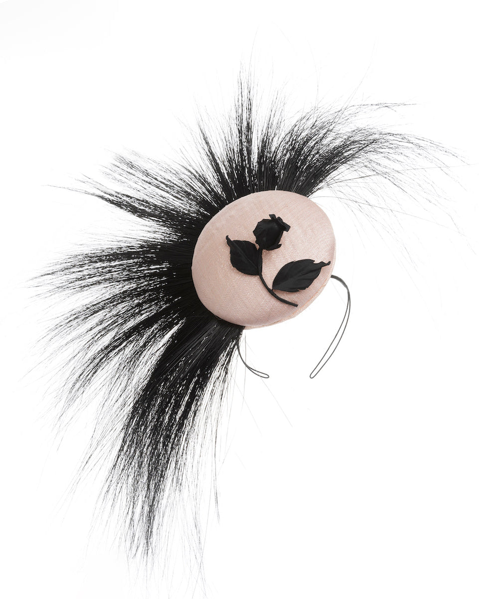 The picture features a pink button-shaped fascinator adorned with strikingly long, burnt-black peacock feathers radiating outward, embellished with a handmade black rose. Designed and made by Merve Bayindir Millinery.
