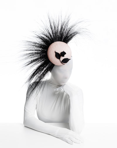 The model is wearing a pink button-shaped fascinator adorned with strikingly long, burnt-black peacock feathers radiating outward, creating a dramatic and eye-catching effect. The fascinator is embellished with an embroidered, handmade black rose, adding a touch of intricate craftsmanship and sophistication.