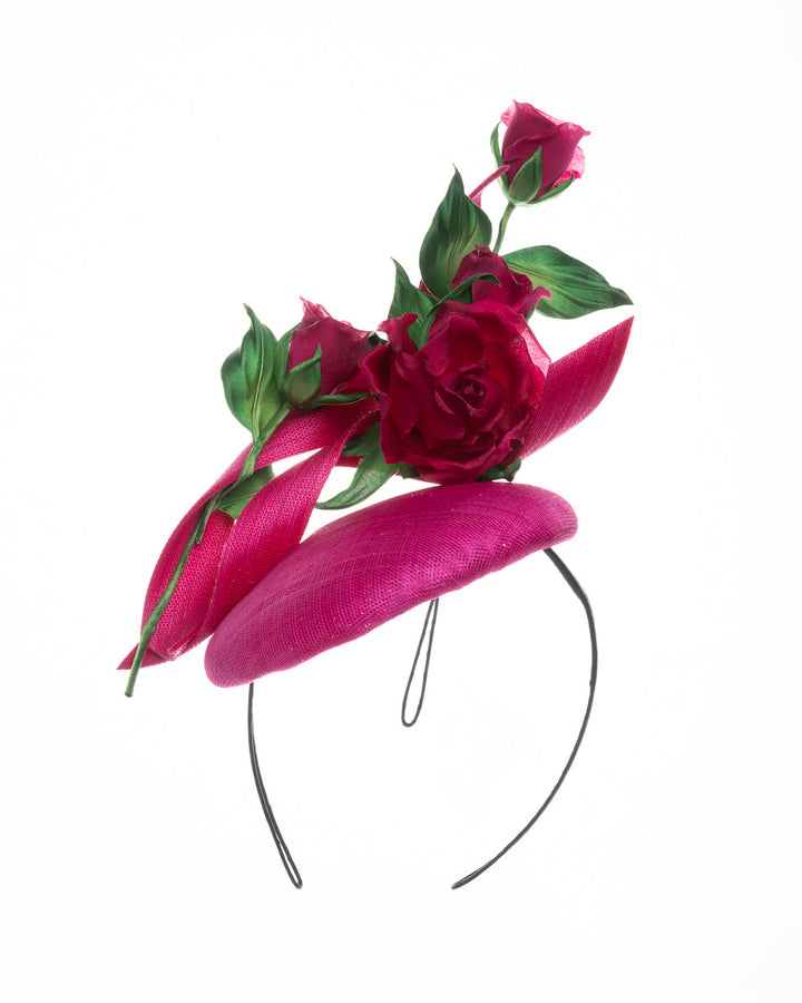 The picture is featuring a hot pink button-style fascinator is decorated with two long, hot pink buntal swirls surrounded by pink roses and rosebuds with green leaves. Designed and made by Merve Bayindir Millinery.
