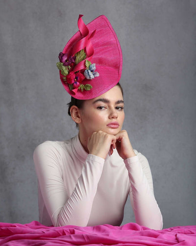 A vibrant pink fascinator with an asymmetrical, leaf-like shape, adorned with a ribbon detail that swirls elegantly. The fascinator is further embellished with a cluster of colorful fabric flowers and green leaves, adding a touch of natural beauty and charm.