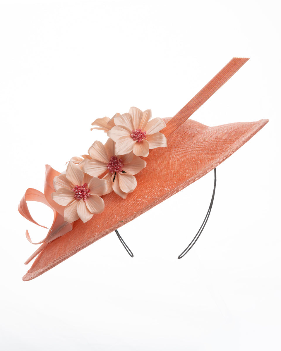 Coral coloured fascinator hat featuring a wide, asymmetrical brim. The hat is adorned with three large, intricately detailed flowers, each with soft, coral coloured petals and pink beaded centres. Designed and made by Merve Bayindir Millinery.
