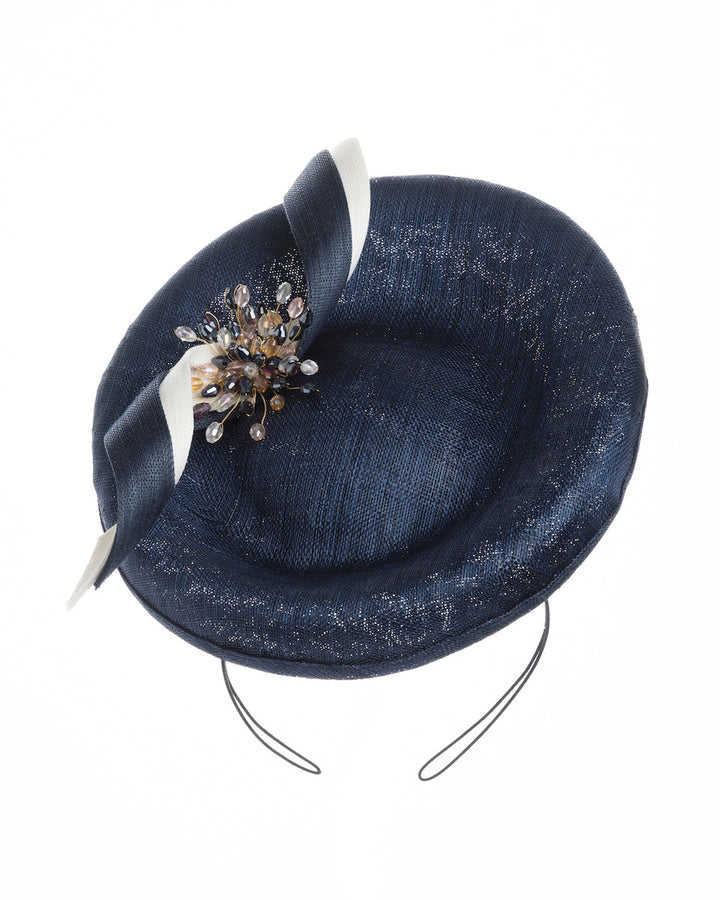 An oval navy fascinator with a structured oval shape sits high on the head. The design is accentuated by graceful navy and white swirls that loop stylishly across the brim, creating a dynamic and eye-catching detail. At the centre of the ribbon, a stunning cluster of crystal beads in various sizes and shades forms a beautiful motif. Designed and made by Merve Bayindir millinery.