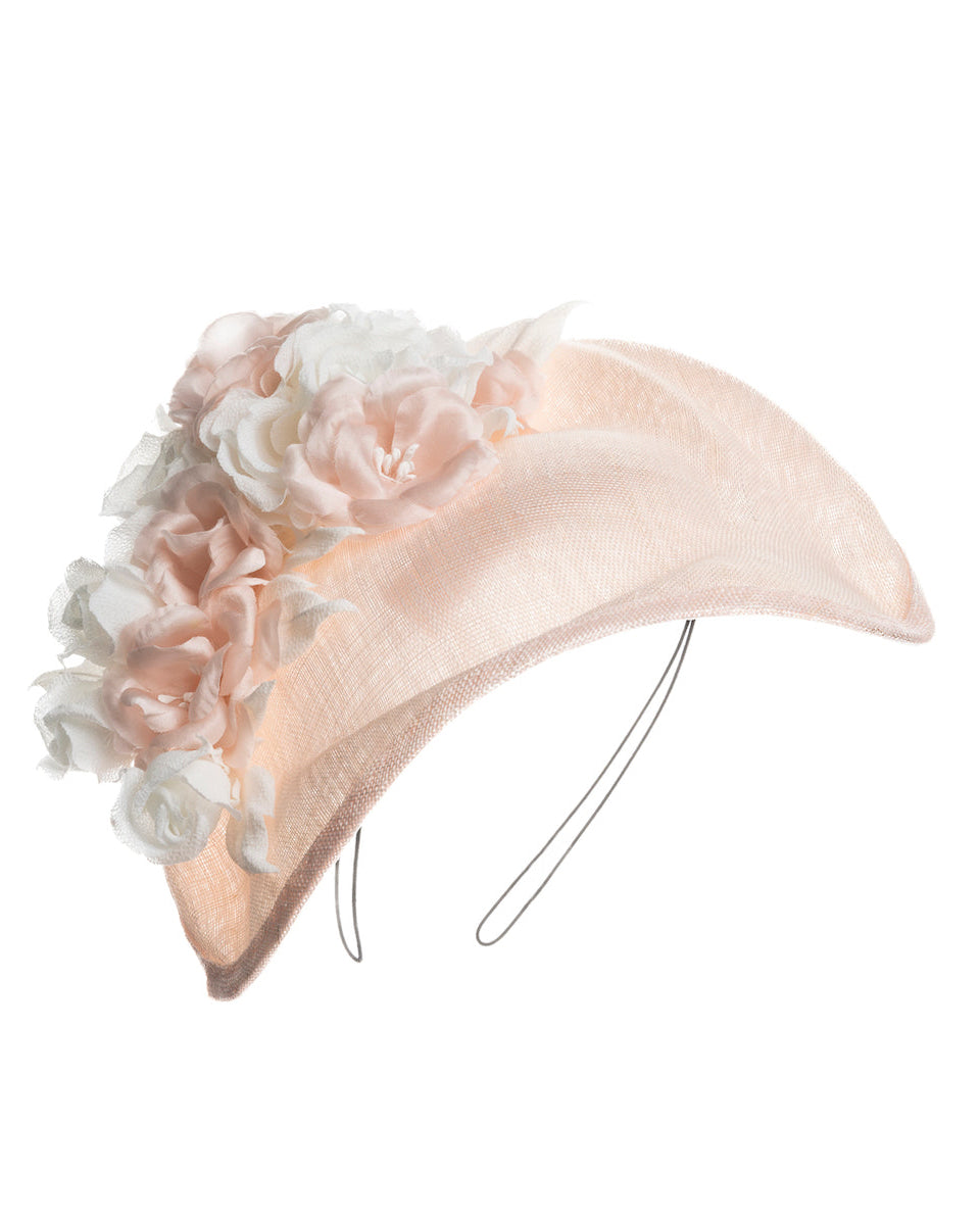 A delicate and elegant pink fascinator hat featuring a soft blush pink fabric with a subtle shimmer. The hat has a structured, crescent shape that arches, providing a flattering and sophisticated silhouette. Adorning the top of the hat is a beautiful arrangement of handmade flowers in soft pink and white hues. Designed and made by Merve Bayindir Millinery.
