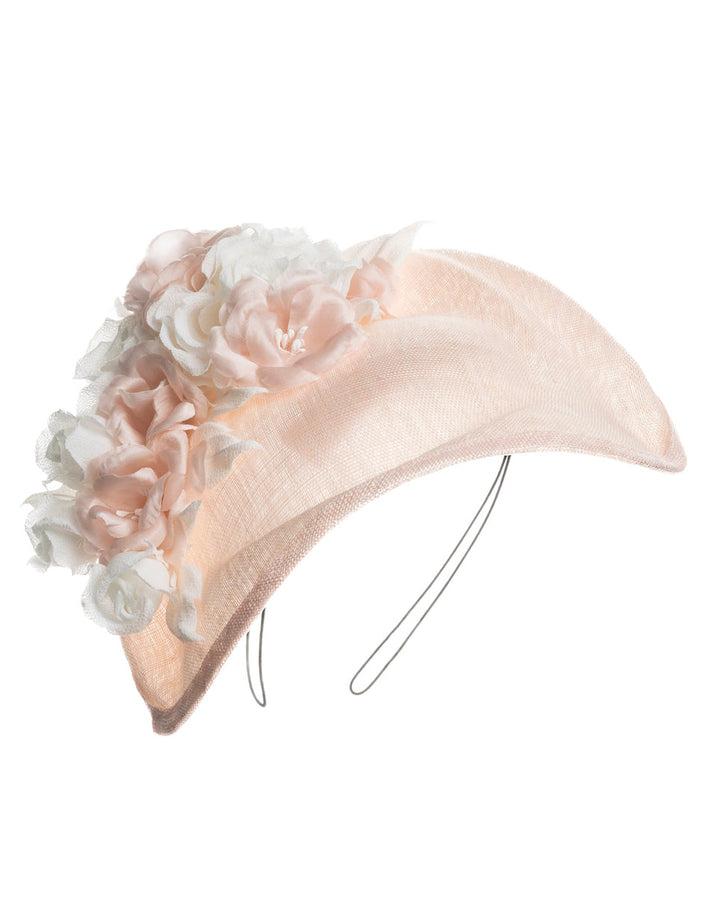 A delicate and elegant pink fascinator hat featuring a soft blush pink fabric with a subtle shimmer. The hat has a structured, crescent shape that arches, providing a flattering and sophisticated silhouette. Adorning the top of the hat is a beautiful arrangement of handmade flowers in soft pink and white hues. Designed and made by Merve Bayindir Millinery.
