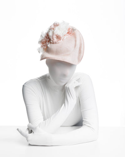 The model is wearing a delicate and elegant pink fascinator hat featuring a soft blush pink fabric with a subtle shimmer. The hat has a structured, crescent shape that arches from back to front, providing a flattering and sophisticated silhouette. Adorning the top of the hat is a beautiful arrangement of handmade flowers in soft pink and white hues,