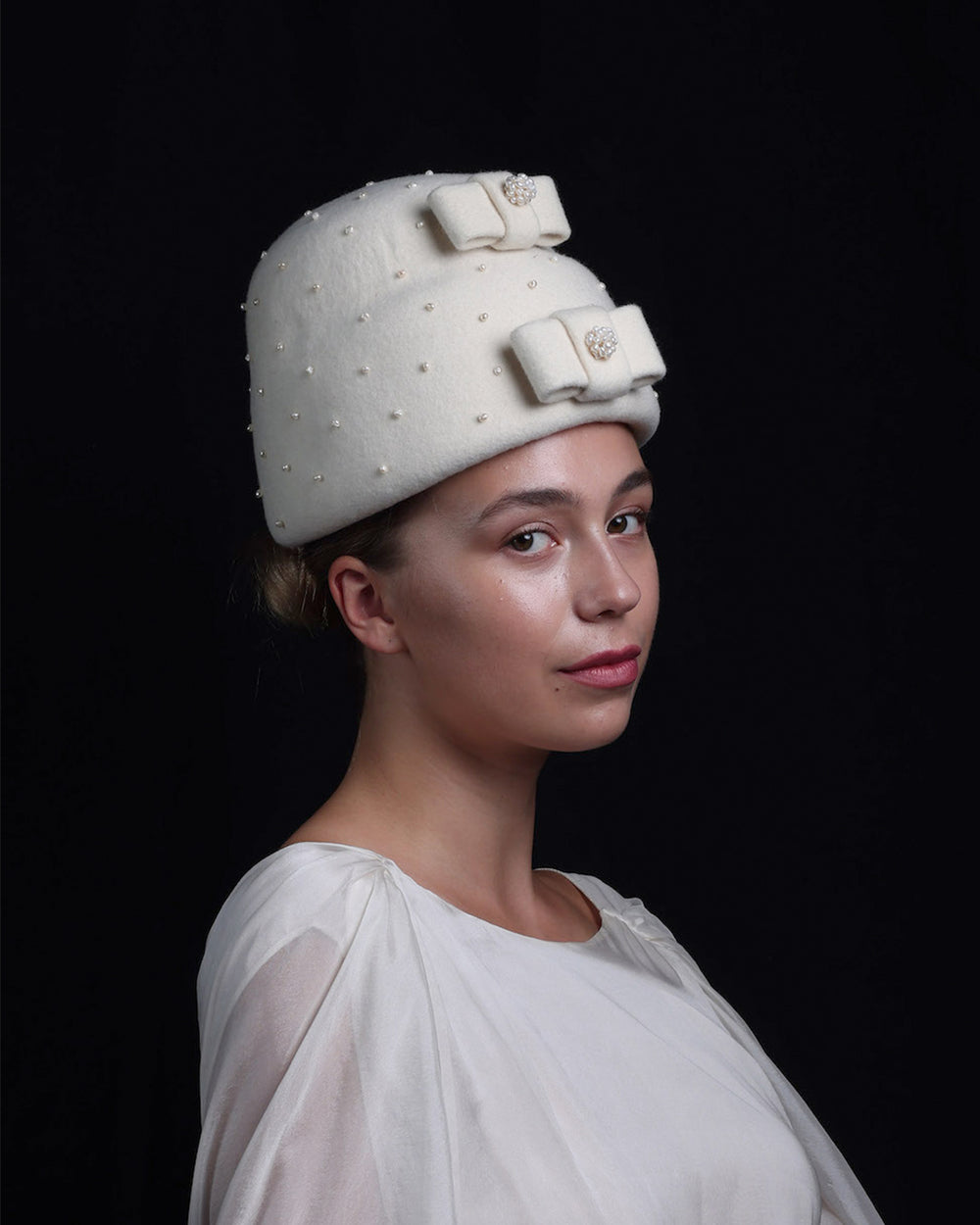 Model is wearing a double layer, tall ivory pillbox hat embellished with pearls.. The hat has ove ivory felt bow on each later, centres adorned with pearl balls. 