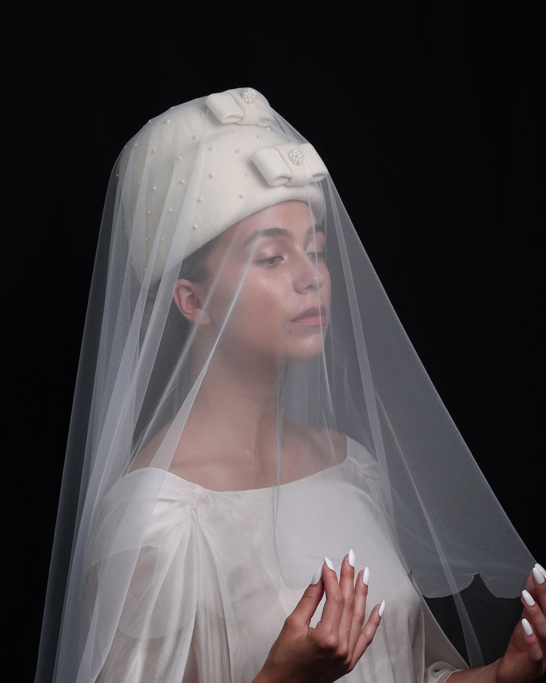Model is wearing a double layer, tall ivory pillbox hat embellished with pearls.. The hat has ove ivory felt bow on each later, centres adorned with pearl balls. The hat is covered with a long bridal veil.