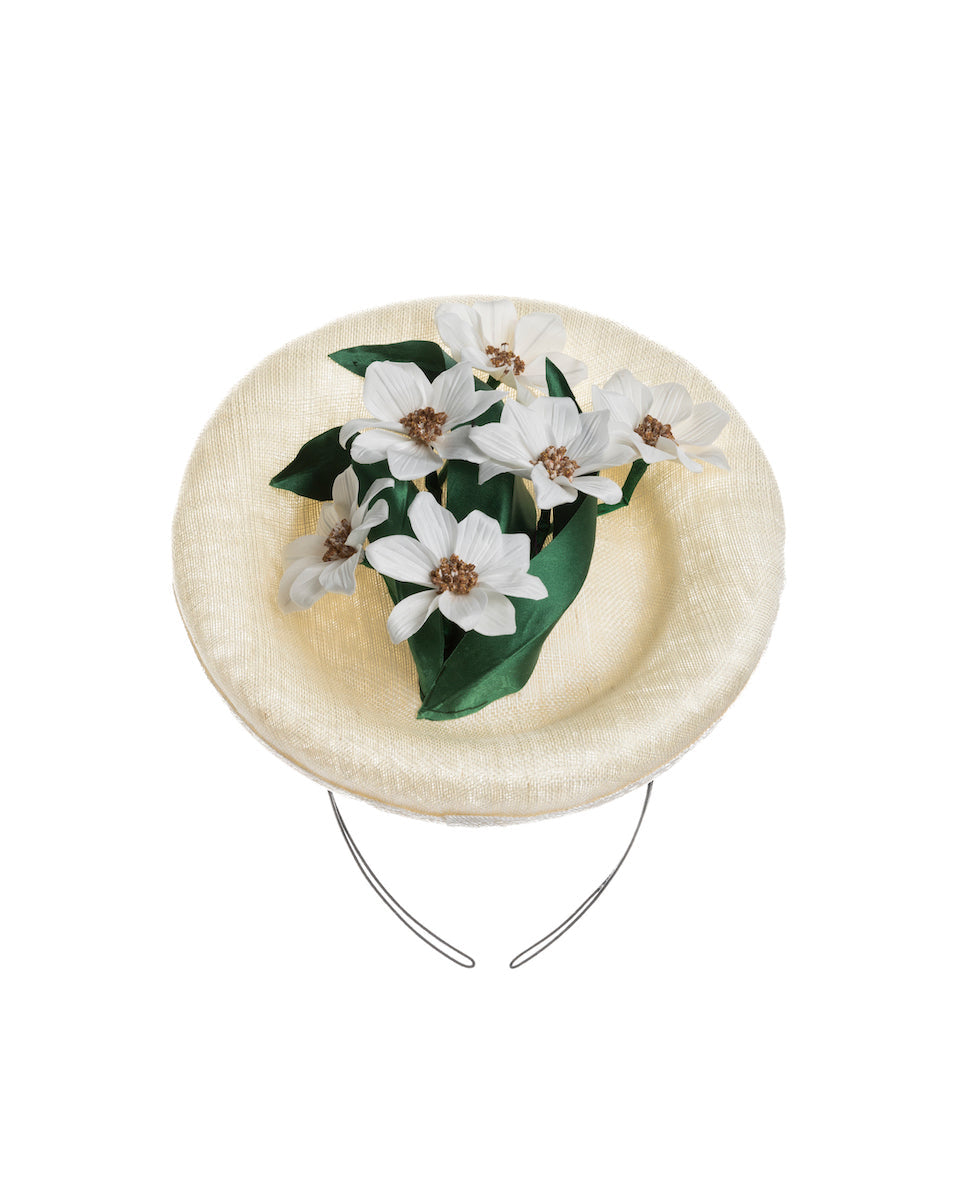 Still life picture of an oval shaped, ivory high saucer fascinator decorated with white silk flowers and green sateen leaves. Designed and Made by Merve Bayindir Millinery.