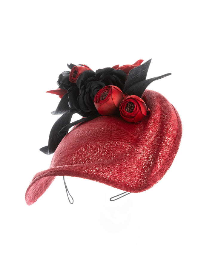 Still life picture of an asymmetric, curved red hat worn to the right side of the head. This headpiece is decorated with a bouquet of black and red crepe roses, black shaded red peonies and black buntal swirls that create a strong contrast and bond between each element. Designed and made by Merve Bayindir Millinery.