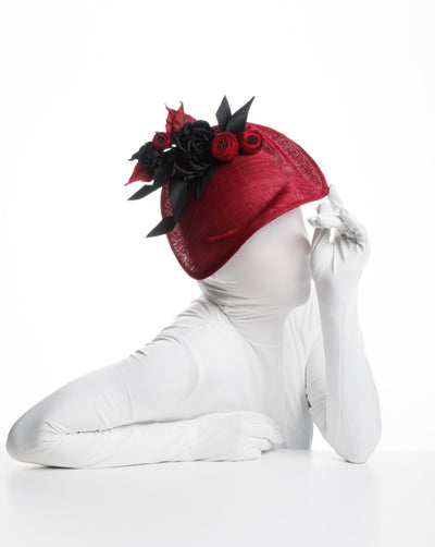 Model is showcasing An asymmetric, curved red hat worn to the right side of the head. This headpiece is decorated with a bouquet of black and red crepe roses, black shaded red peonies and black buntal swirls that create a strong contrast and bond between each element.