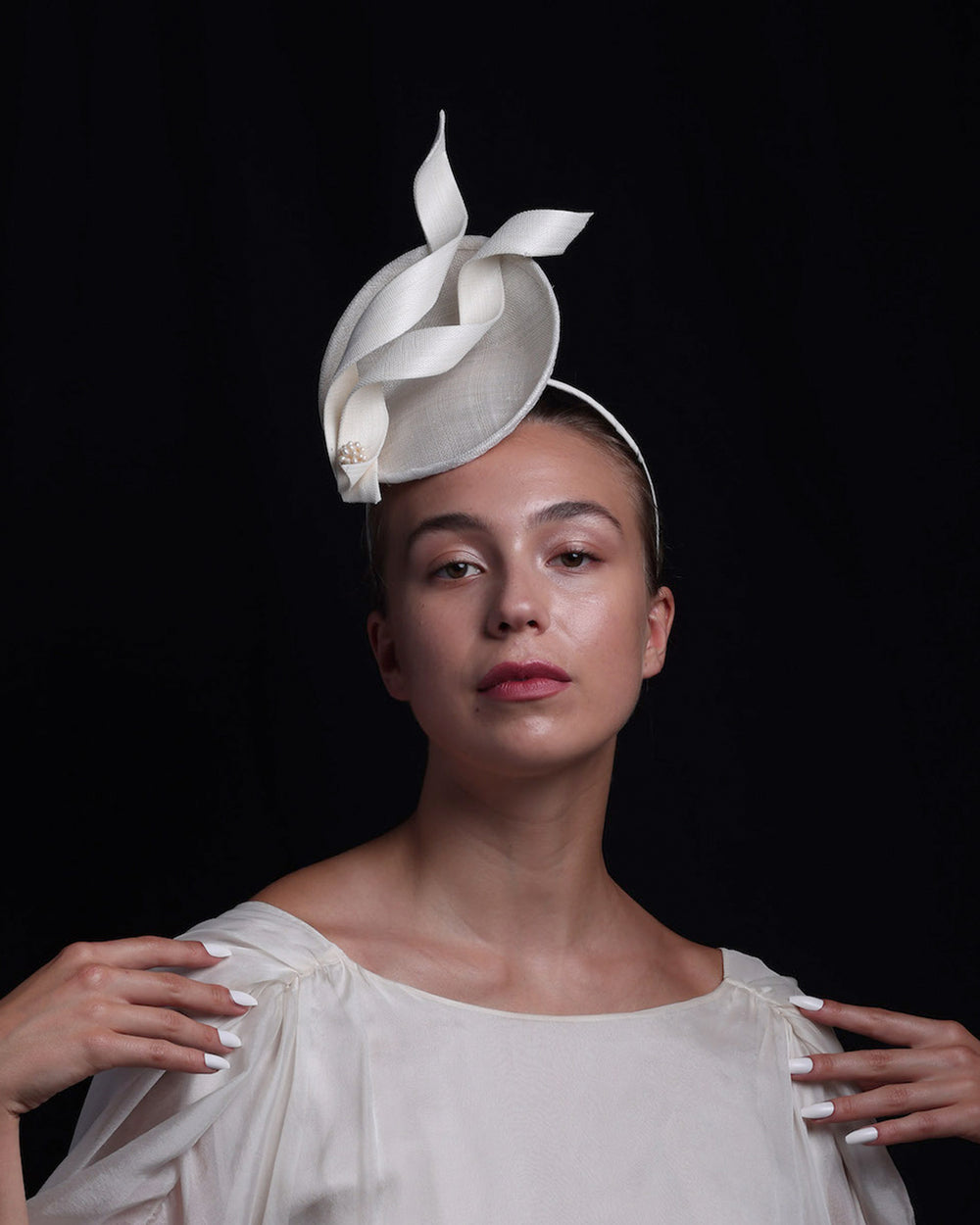 Model is wearing an off-white, mini saucer style fascinator decorated with two buntal swirls and a mother of pearl ball.