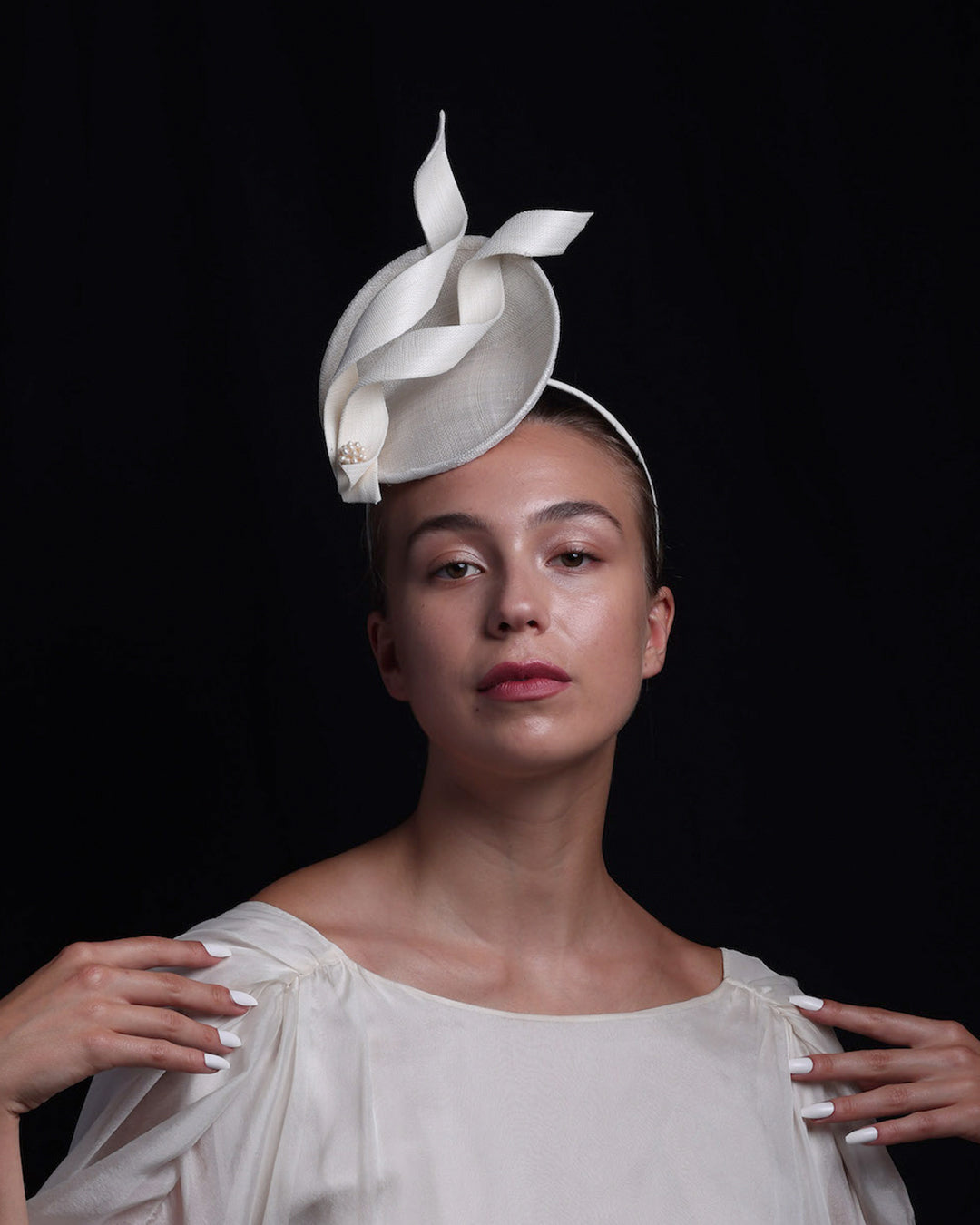 Model is wearing an off-white, mini saucer style fascinator decorated with two buntal swirls and a mother of pearl ball.