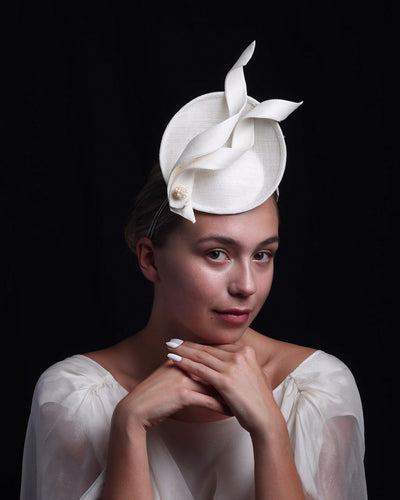 Model is wearing an off-white, mini saucer style fascinator decorated with two buntal swirls and a mother of pearl ball.