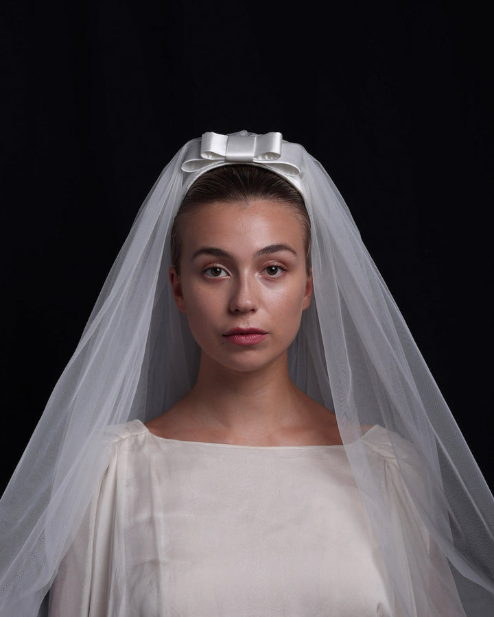 Model is wearing a short white sateen headband decorated with a double layered sateen bow and completed with a two-pieced ghost tulle veil.