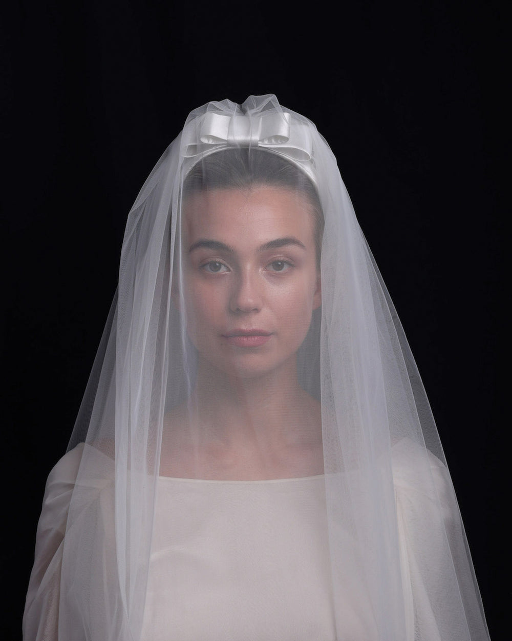 Model is wearing a short white sateen headband decorated with a double layered sateen bow and completed with a two-pieced ghost tulle veil covering the brides face
