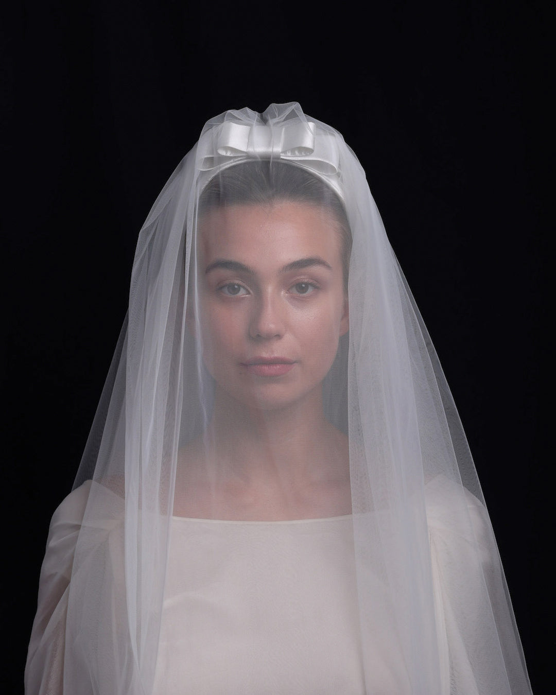 Model is wearing a short white sateen headband decorated with a double layered sateen bow and completed with a two-pieced ghost tulle veil covering the brides face