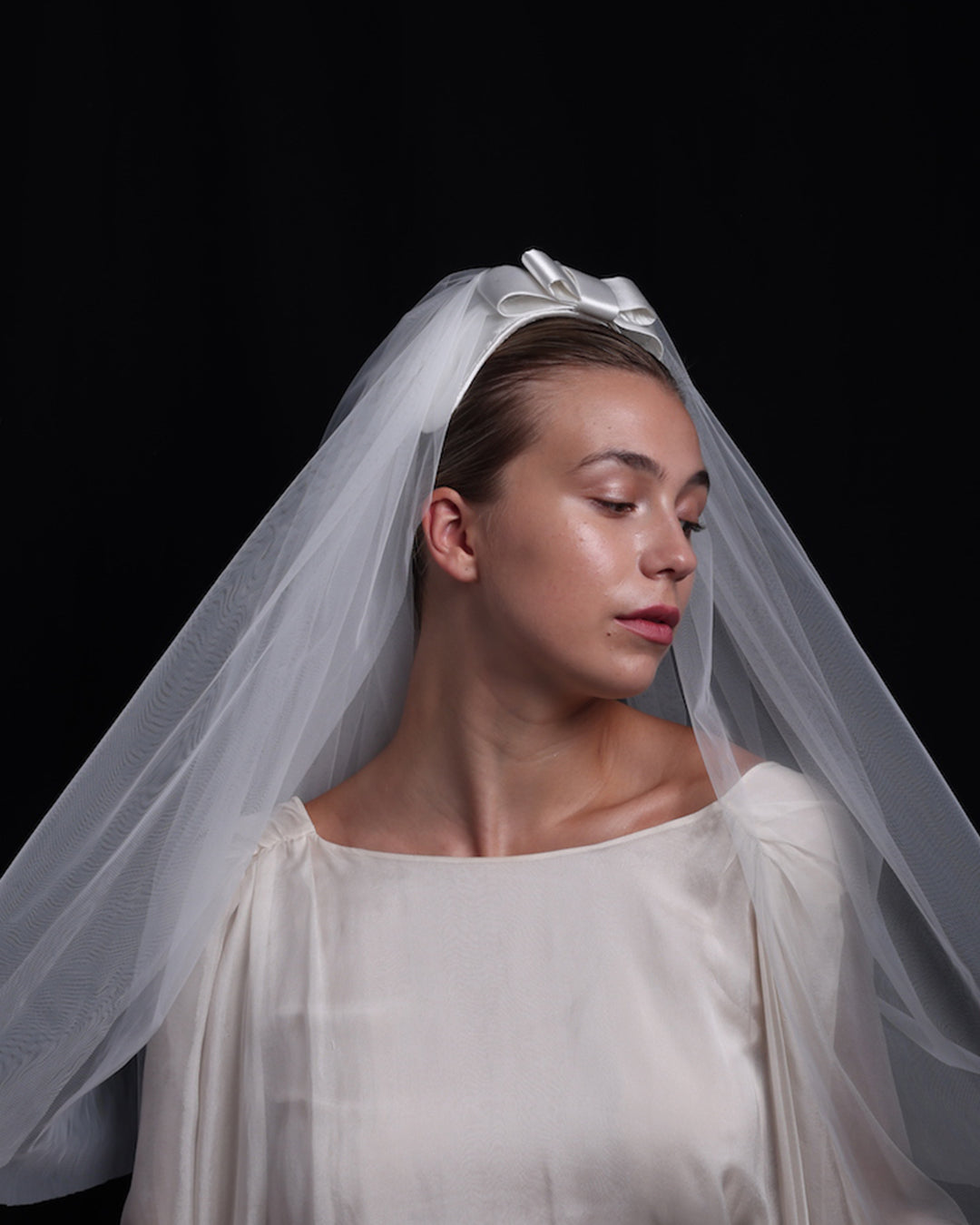 Model is showcasing  a short white sateen headband decorated with a double layered sateen bow and completed with a two-pieced ghost tulle veil.