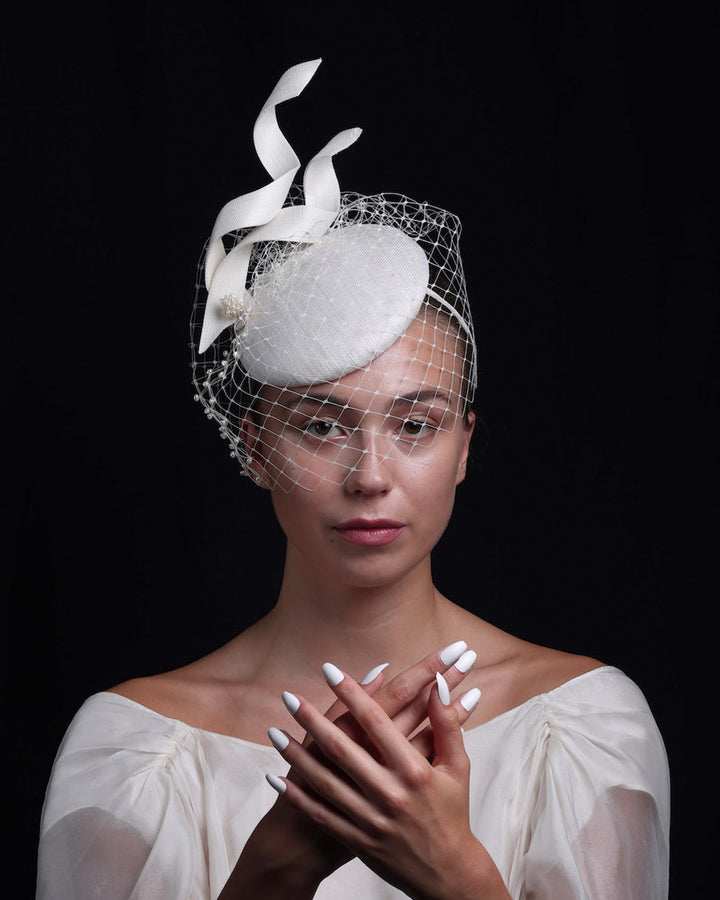 Model is showcasing a white button shaped bridal fascinator decorated with two buntal swirls and rounded with mother of pearl embroidered veil.