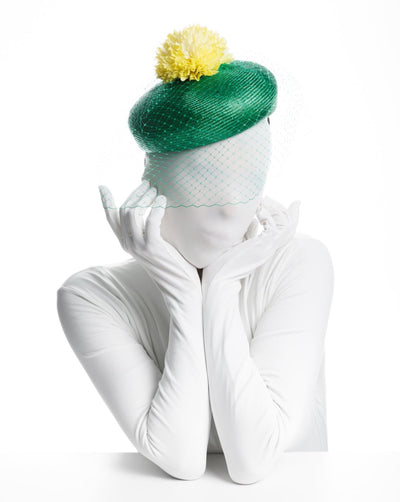 Model is wearing a Emerald green summer beret tilted to the right side of the head. This vibrant headpiece is rounded with a delicate emerald green veil and completed with a bright yellow pompom flower on top.