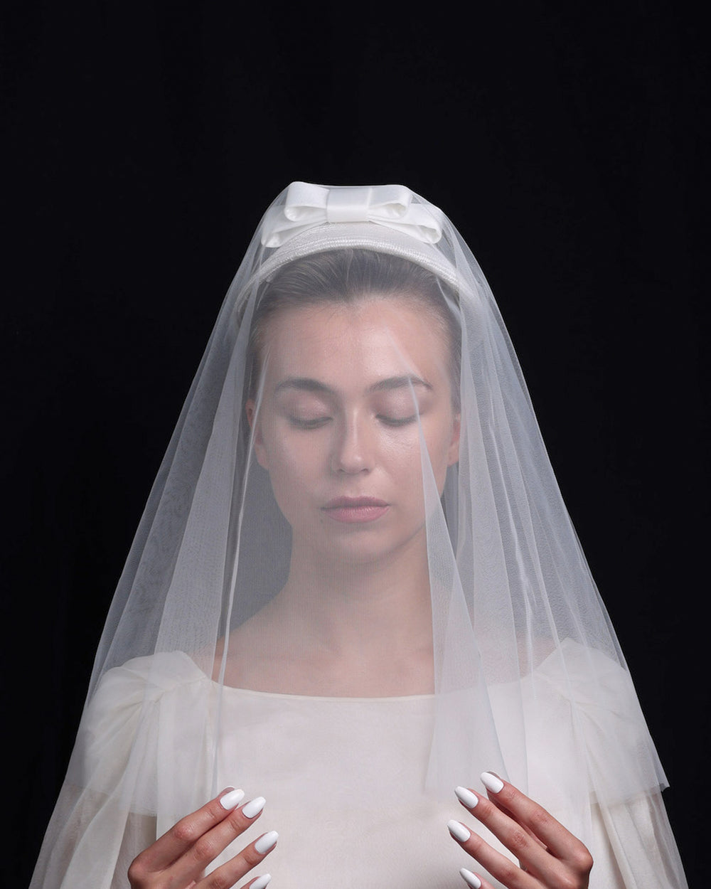 A front look of White curved, parasisal, pillbox style bridal headpiece decorated with a double layered sateen bow and completed with a shoulder length, circle ghost tulle veil..