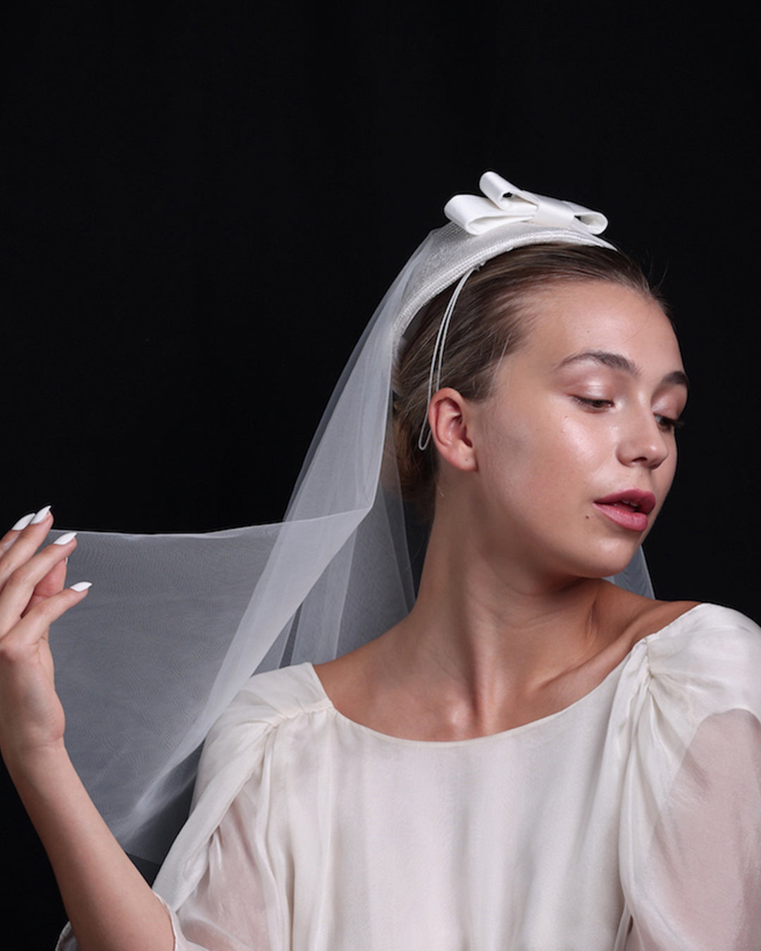 A side look of a White curved, parasisal, pillbox style bridal headpiece decorated with a double layered sateen bow and completed with a shoulder length, circle ghost tulle veil.