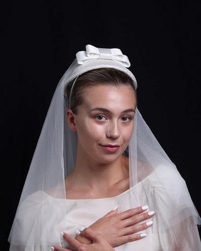 A front look at White curved, parasisal, pillbox style bridal headpiece decorated with a double layered sateen bow and completed with a shoulder length, circle ghost tulle veil. The veil is thrown back of the hat.