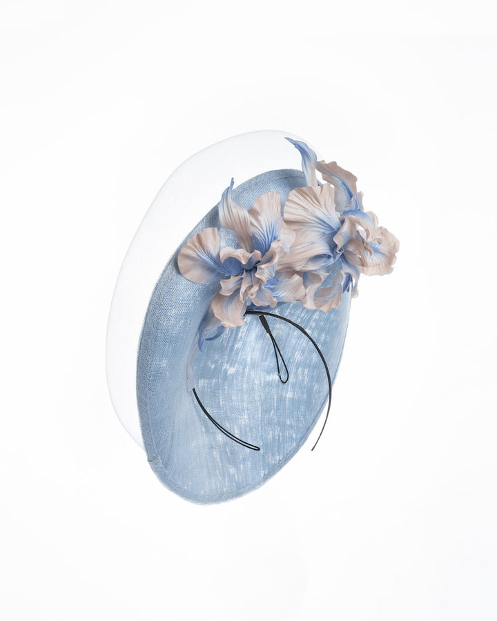 A medium sized, side saucer shaped light blue headpiece that blends different styles into one. Almost vertical construction of this splendid design softens with a crinoline curve on the left side and features light pink and light blue shaded irises underneath. Designed and made by Merve Bayindir Millinery