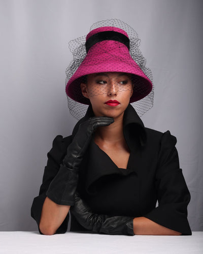 Model is featuring an oversize pink felt bucket hat decorated with veil, a wide velvet belt and a large pearl bead in the back.