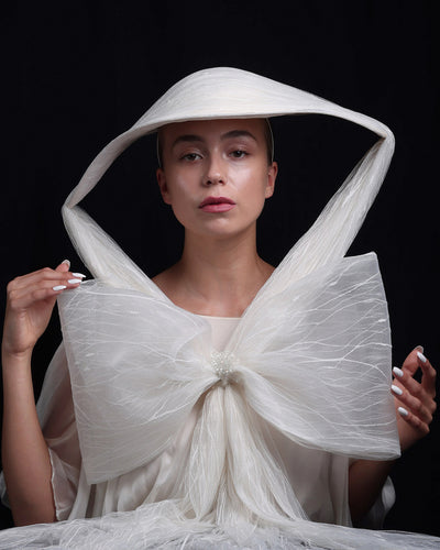 Model is showcasing an asymmetric wide brim hat draped with a glittered and embroidered tulle from the front. The voluminous tulle is tied with a large bow and decorated with a crystal button on the front.