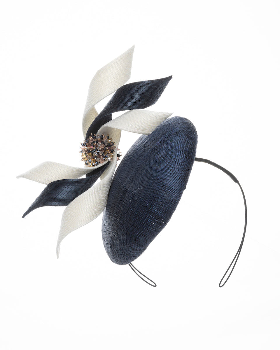 A navy, button shaped fascinator worn to the right side of the head. This chic headpiece is decorated with navy and white buntal swirls joined by multi coloured crystal beads, creating a flower-like silhouette. Designed and made by Merve Bayindir millinery.