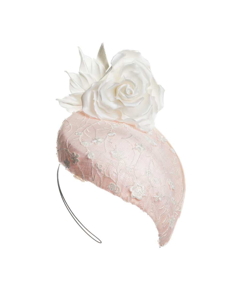 Light pink, percher style sinamay hat that curves on the left side of the head, covered with flower detailed white lace. This pink fascinator features a large, white silk satin rose that enhances the visual impact. Designed and made by Merve Bayindir Millinery.
