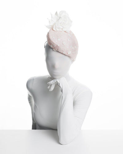 The model is showcasing the front view of the light pink, percher style sinamay hat that curves on the left side of the head, covered with flower detailed white lace. This pink fascinator features a large, white silk satin rose that enhances the visual impact.