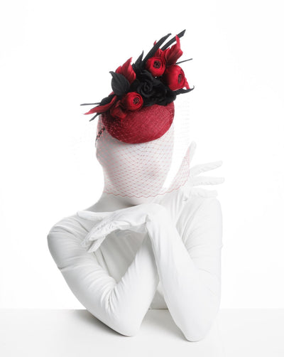 Model is showcasing the right side of a red button shaped fascinator worn to the centre to the right side of the head. This fascinator hat is decorated with a bouquet of black crepe roses, black shaded red peonies and black buntal swirls, and, rounded with a delicate red veil that covers the face.