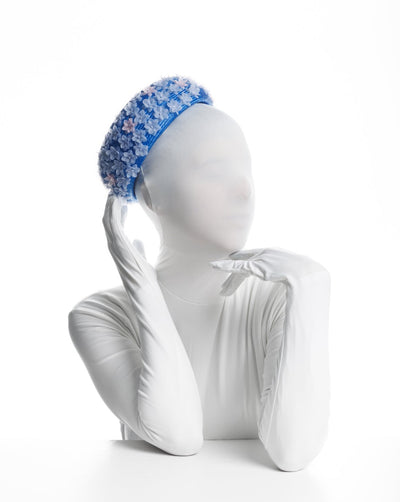 Model is showcasing the side of a deep blue, pillbox style hat worn to the back of the head. Decorated with mini blue and pink organza flowers.