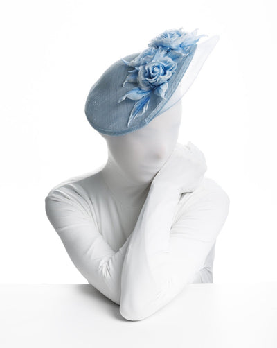 Model is showcasing a front view of a light blue, asymmetric sinamay hat decorated with a shaded blue rose bouquet on the top centre and a light blue, crinoline curve on the left edge. Leaving the left side of the head fully exposed.