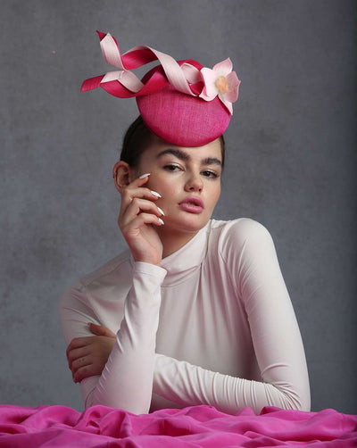 Model is wearing a high fashion button shaped pink fascinator decorate with jinsin swirl ribbons and a light pink orchid.