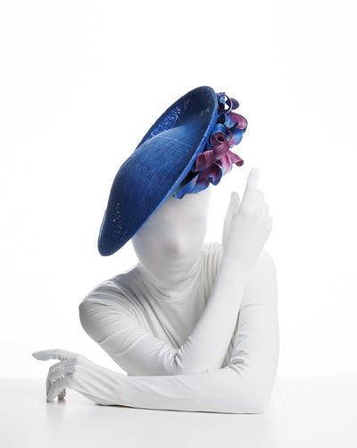 The model is showcasing the front of the a deep blue, large side saucer hat with a twist. While calming as blue skies on the left, right side of this wide brim saucer hat has a very playful with purple and deep blue leaves that swirl under the curved brim.