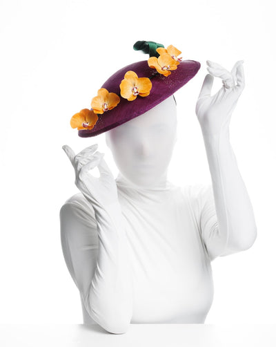 Model is presenting a front view of  a deep purple, round sinamay saucer hat decorated with orange orchids and a green swirl leaf.