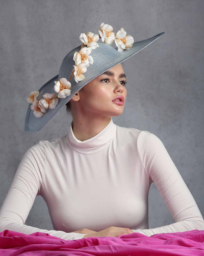 The model is showcasing an asymmetric wide brim blue hat decorated with custom made American sateen flowers from one edge to the other.