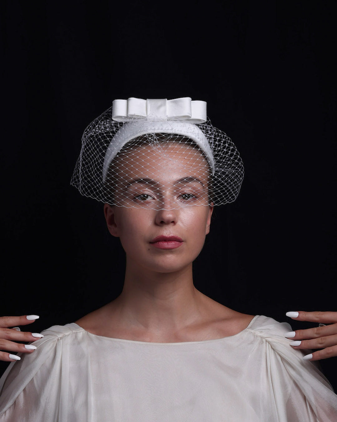 Model is showcasing the front view of sateen bridal headband decorated with an upright, double layered sateen bow. This minimalistic headpiece is completed with a delicate, round hat veil.