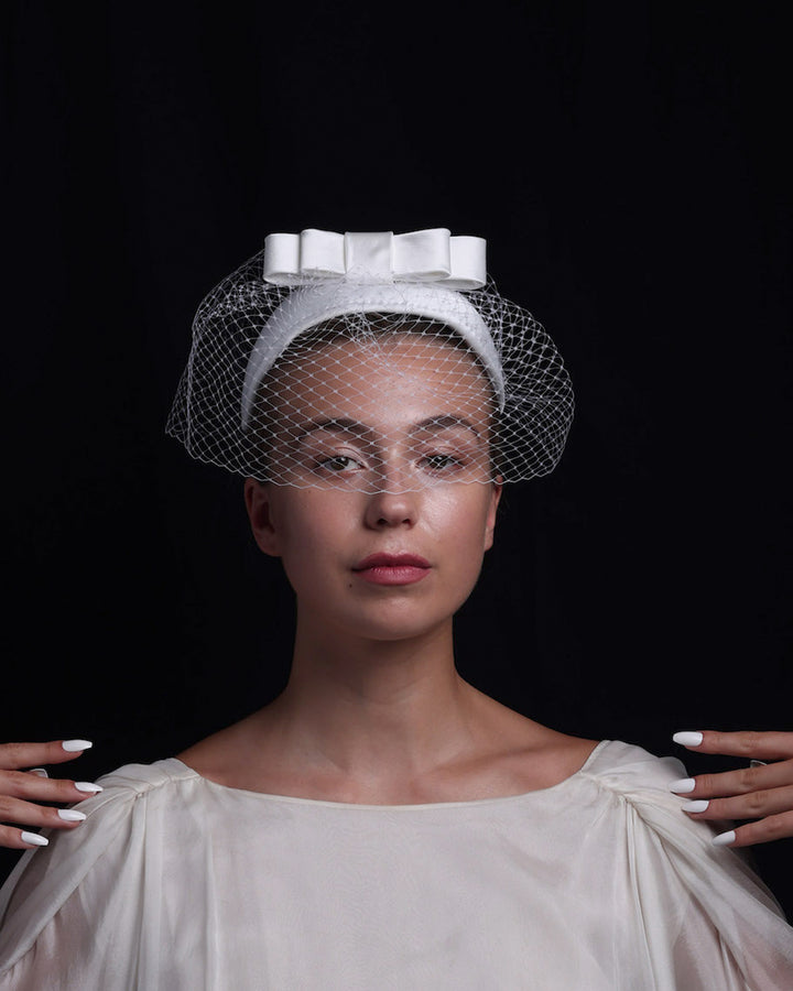 Model is showcasing the front view of sateen bridal headband decorated with an upright, double layered sateen bow. This minimalistic headpiece is completed with a delicate, round hat veil.