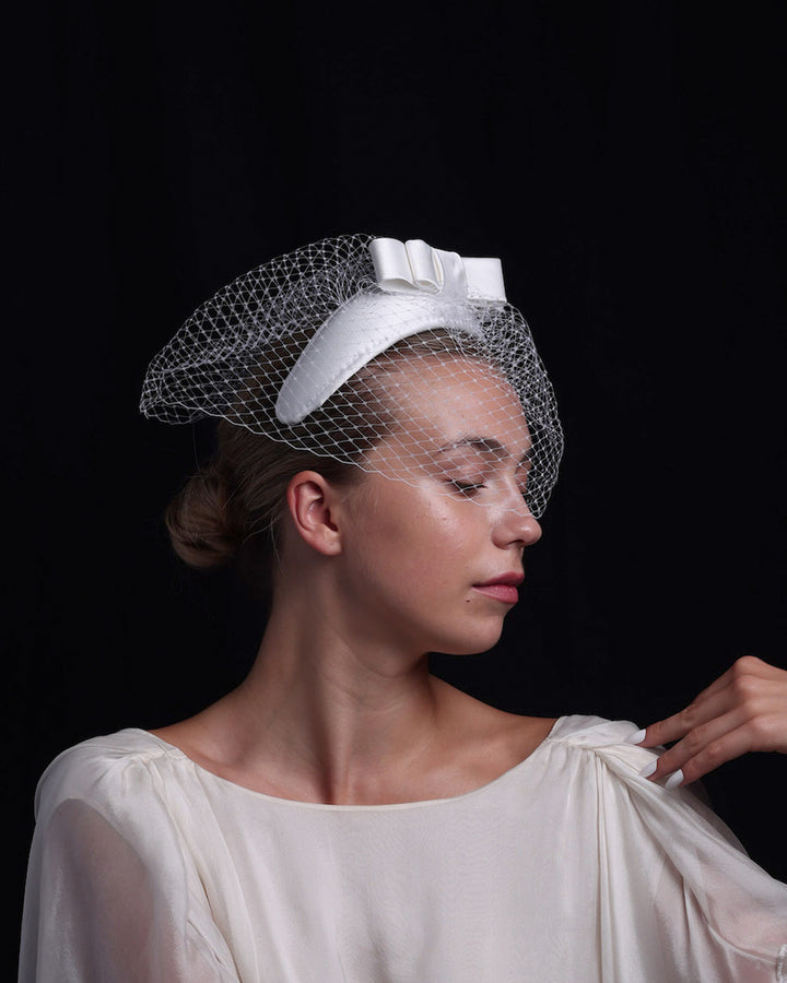 Model is showcasing the side view of a sateen bridal headband decorated with an upright, double layered sateen bow. This minimalistic headpiece is completed with a delicate, round hat veil.