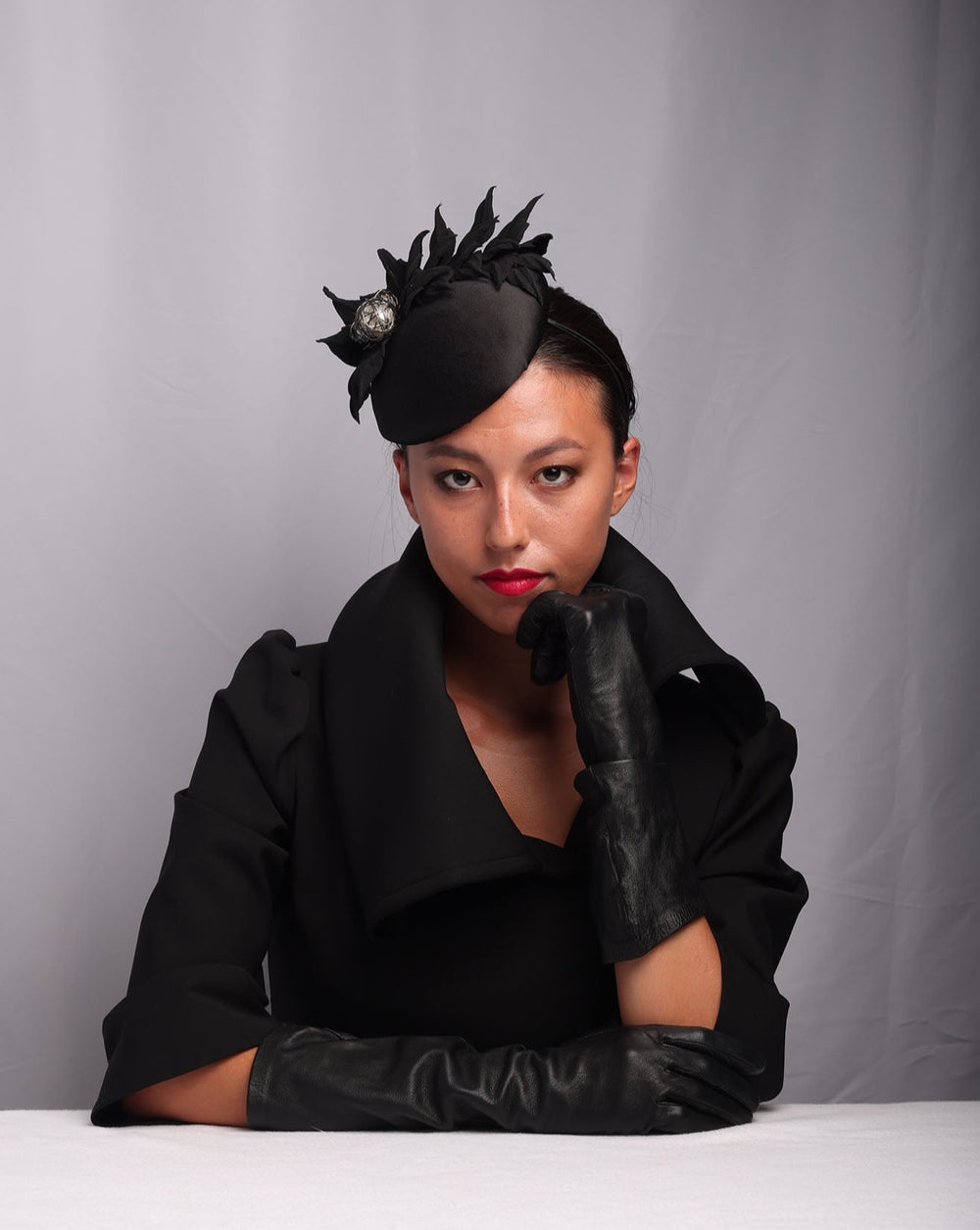 Model is showcasing The button style sateen based fascinator decorated with crepe leafs and centred with wire embellished plastic large pearls.