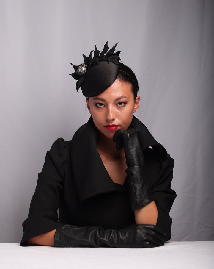 Model is showcasing The button style sateen based fascinator decorated with crepe leafs and centred with wire embellished plastic large pearls.
