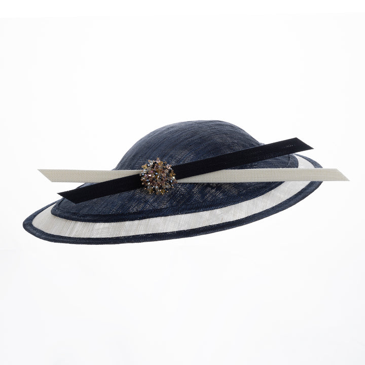 Still life picture of multi layered tear drop shaped saucer in navy and white. This wide brim hat is decorated with navy and white buntal stripes and multi coloured crystal embellishments on the front. Designed and made by Merve Bayindir millinery.