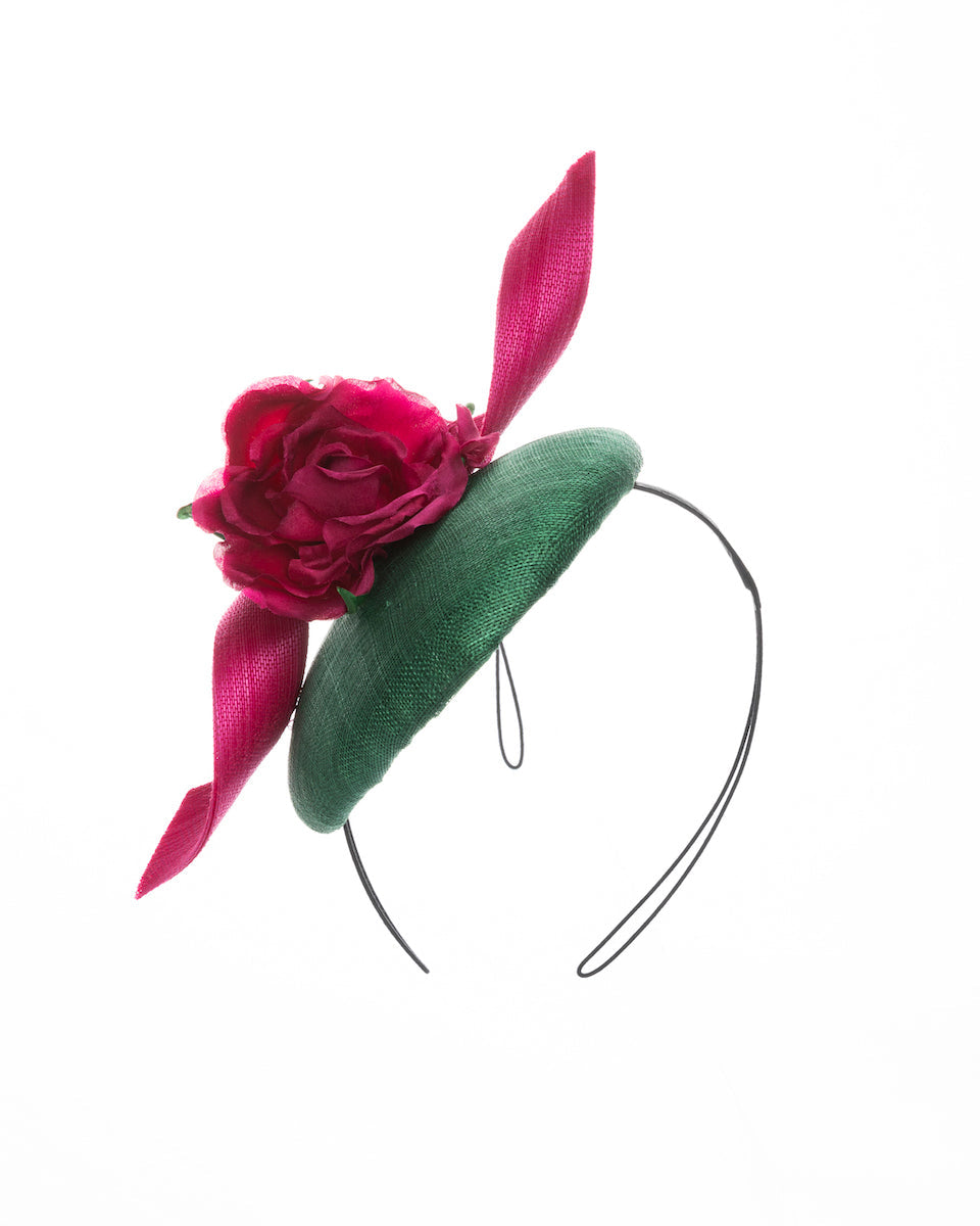 Button-shaped green fascinator with a pink jinsin swirl crossing the base, the centre of the swirls adorned with a hot pink rose. The fascinator sits on the right front side of the head. Designed and made by Merve Bayindir millinery.